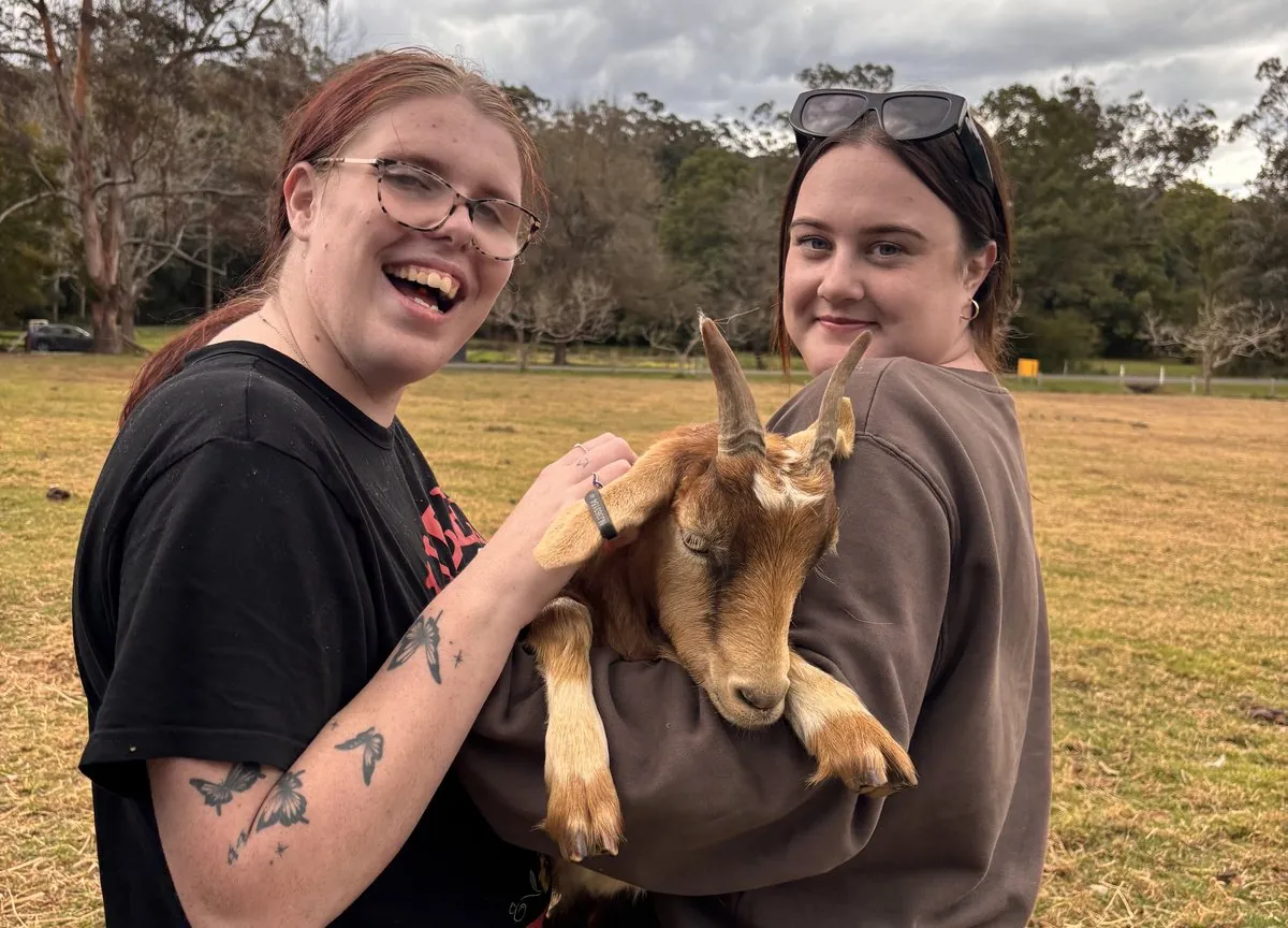 A client and her support worker sharing a laugh while cuddling a goat on a community farm visit